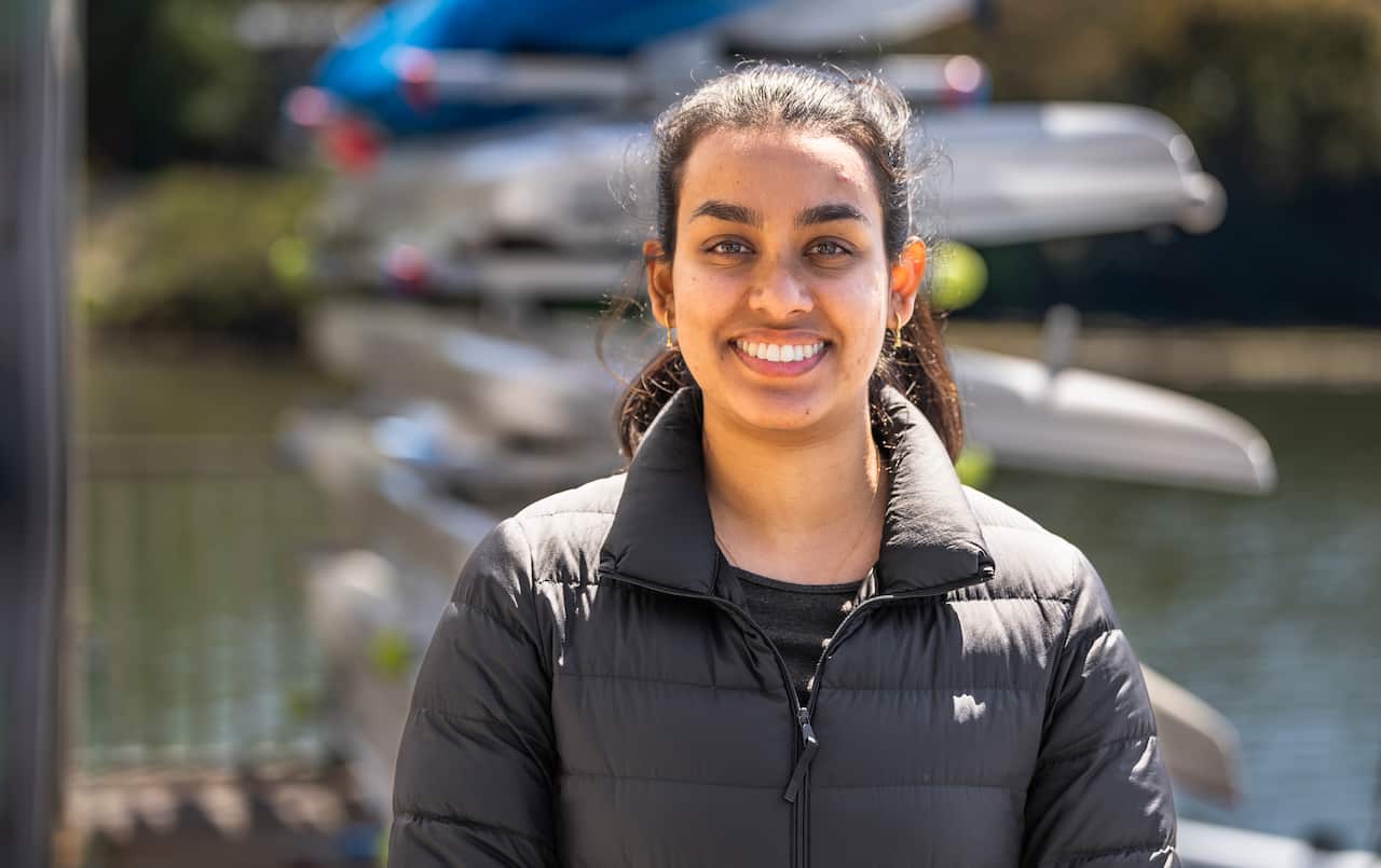 A young woman smiles at the camera in front of rowing boats.