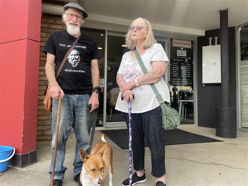 An elderly couple, with a dog, outside a building.