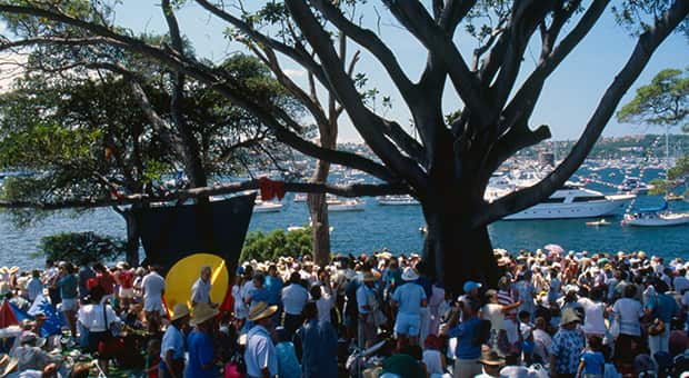 A hill, dominated by a large fig tree, is covered with people wearing white clothes. A large aboriginal flag hangs from one of the branches. Immediately behind is Sydney harbour, covered with white boats.