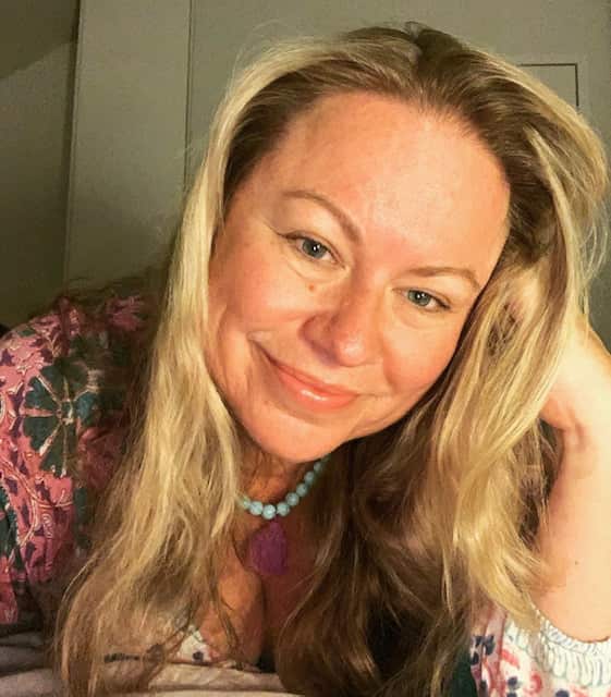 A woman with long blonde hair leans on her hand and smiles at the camera.