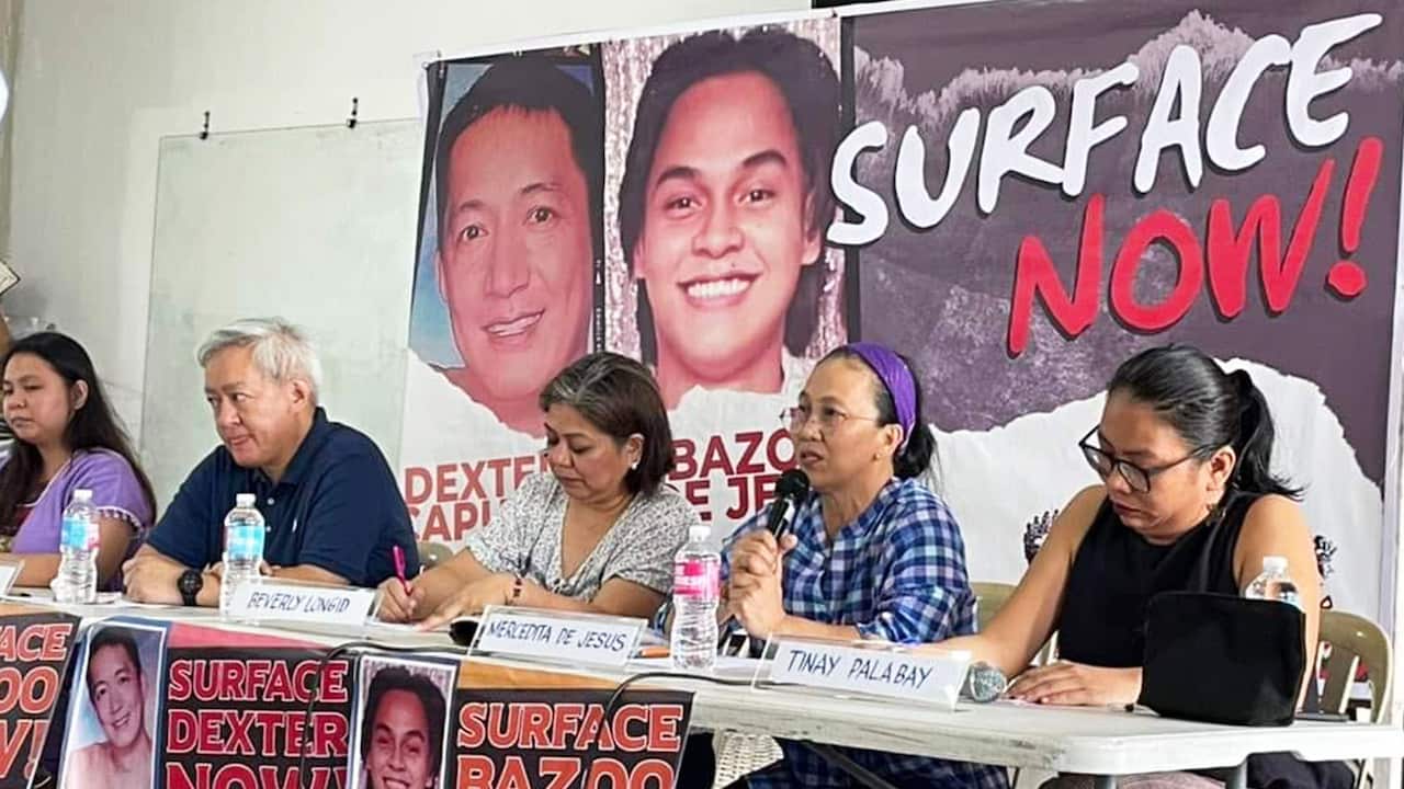 At a press conference, five people —a grey-haired man and four women—sit along a long table adorned with posters reading 'Surface now!' and depicting portraits of two Filipino men, Dexter Capuyan and Bazoo De Jesus. The woman second from the right is wearing a blue plaid shirt and a purple hairband and is speaking into a microphone, holding it with both hands. 