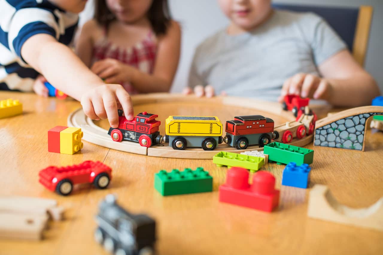 The torsos and arms of three young children playing with trains on wooden track at a table.