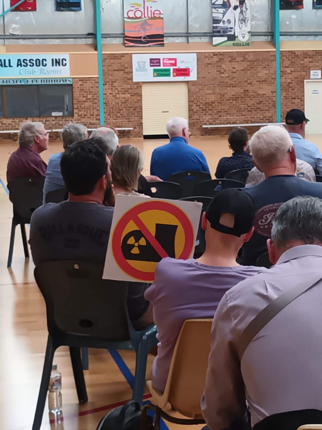 An audience sits on folding chairs in a sports hall. One of them has a sign on the back of his chair with a line through an image of a nuclear reactor and radiation symbol