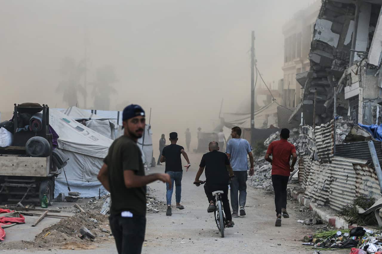A group of men flee down a dirt road, which is covered in rubble and dust. The man in the foreground looks back over his shoulder while the others run away from the camera, with one man riding a bike and another man's face obscured by the dust.
