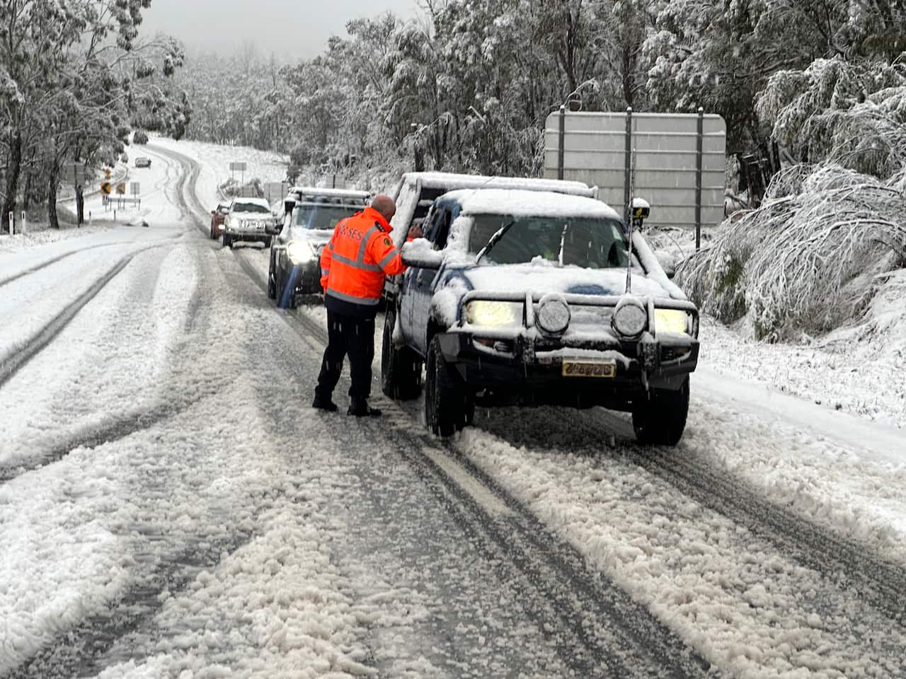 A rescue worker is speaking to the driver of a car on a snowy road. There's a row of cars behind the first vehicle.