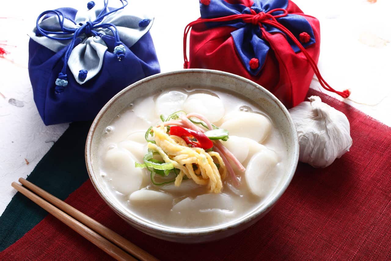 A bowl of rice cake soup surrounded by a garlic bulb, two pouches in blue and red, and chopsticks placed on a red tablecloth.