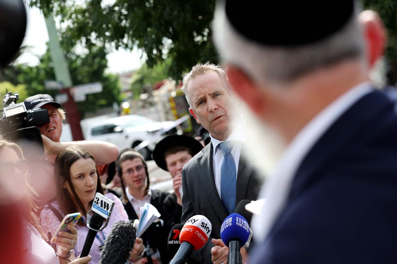 A man in a suit with an angry expression stands in front of microphones held by reporters, staring at an elderly man wearing a kippah.