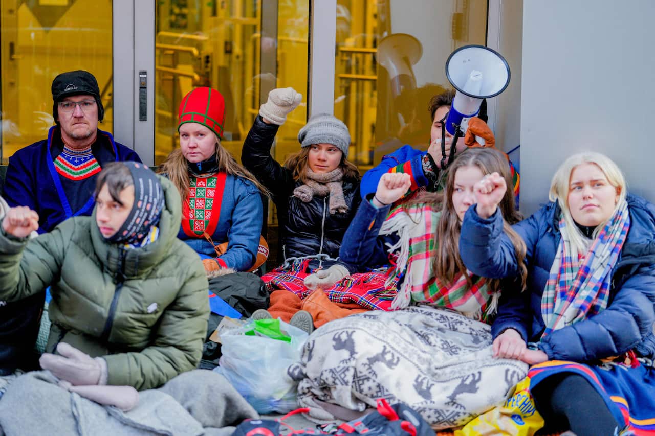 A mixed group of young men and women in warm winter clothes sitting on the ground outside of a building raising their right fists in protest