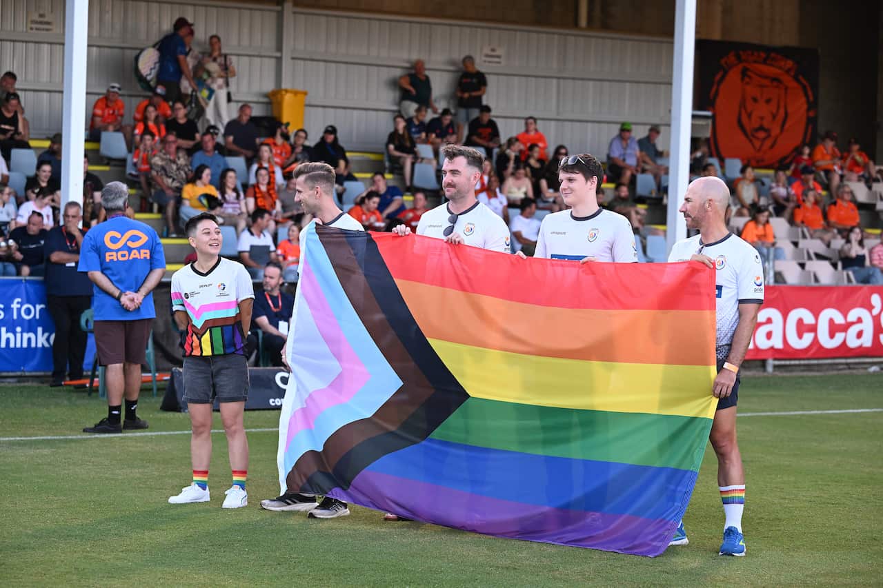 An LGBTIQ+ flag is being held up by three male football players on a football field