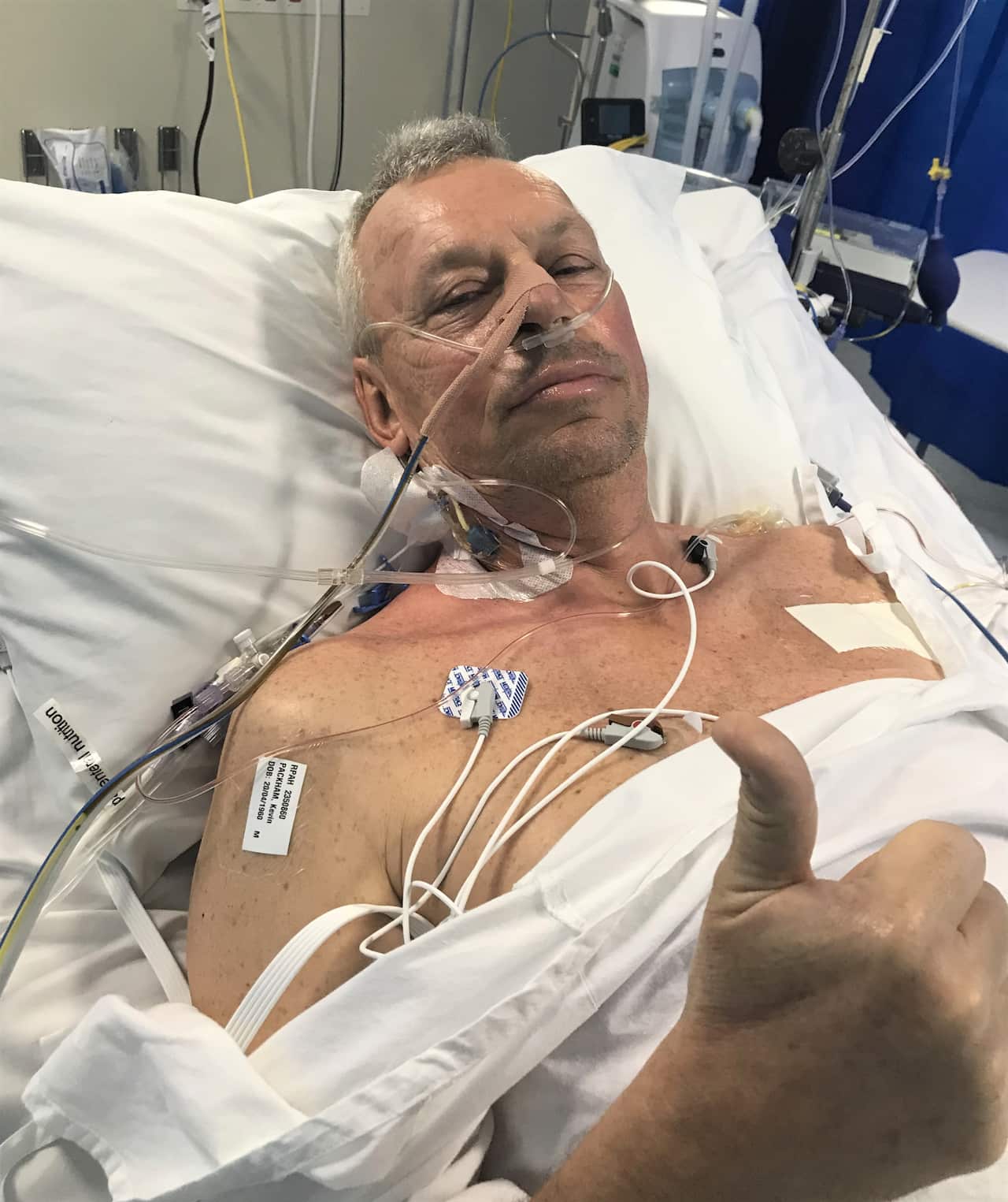 A man in a hospital bed giving a thumbs-up sign. He has wires attached to his chest and a tube attached to his nose.