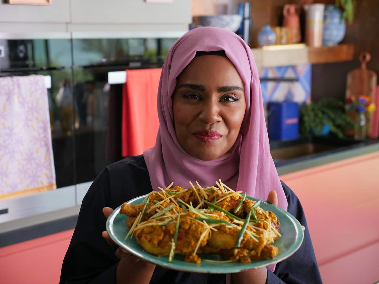 A woman in a pink headscarf holds up a green plate of coated chicken pieces. 