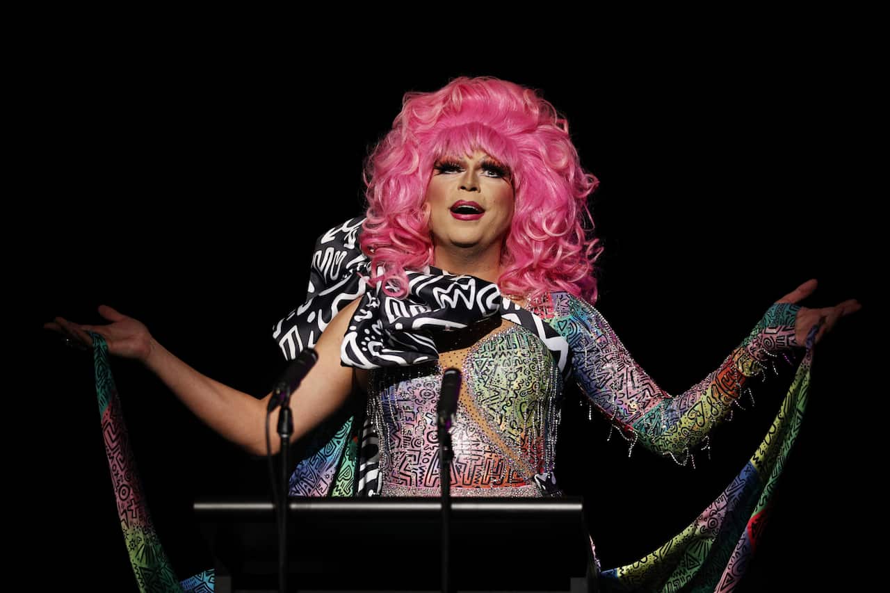 Ben graetz in his drag persona Miss Ellaneous stands at a lectern, wearing a large pink wig and a first nations-design patterned dress, with a big black and white bow on his shoulder. His arms are raised and he smiles upwards at the lights