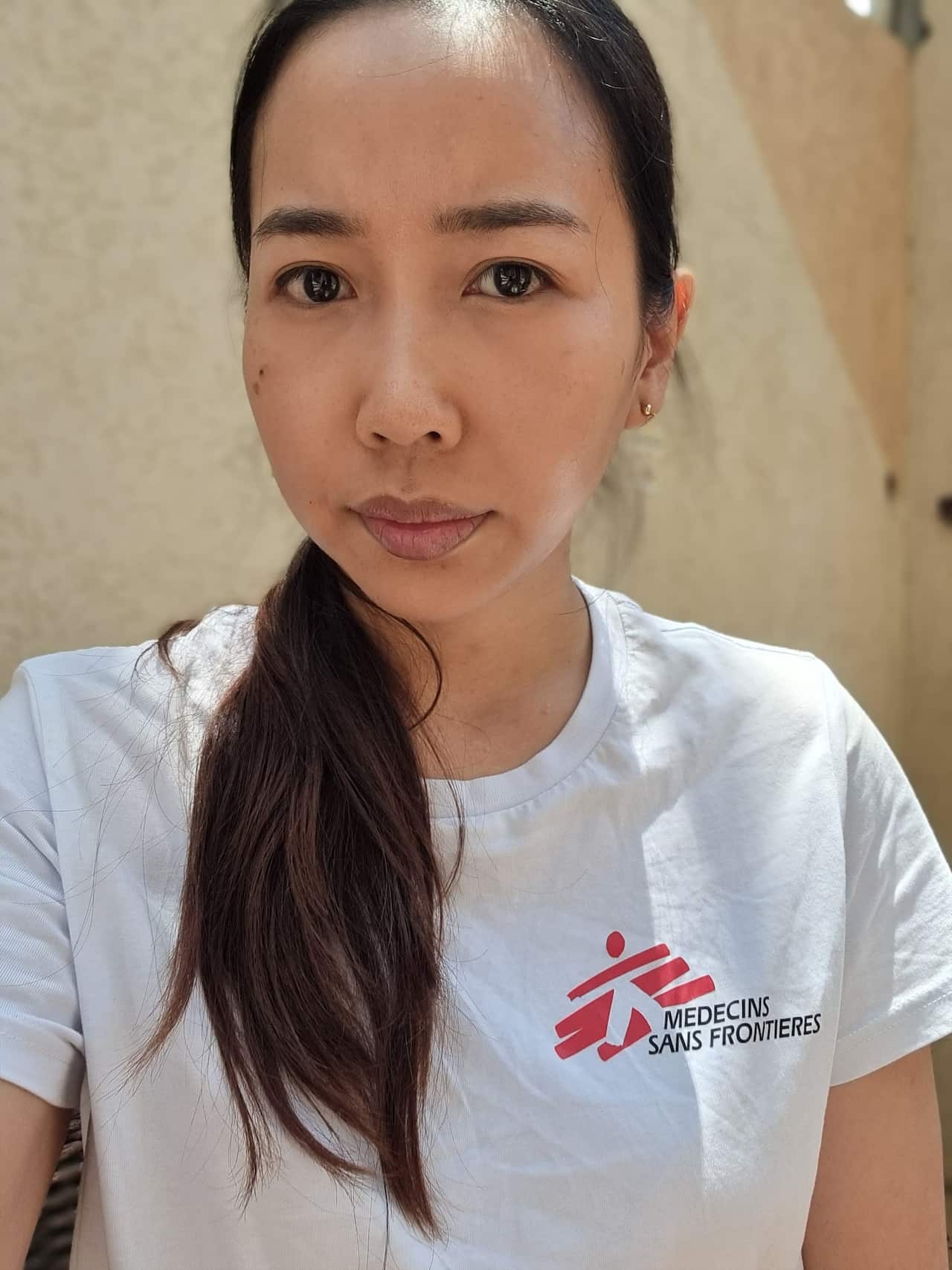 A doctor in a white shirt with the "Medicins Sans Frontieres" logo and long black hair in a ponytail. 