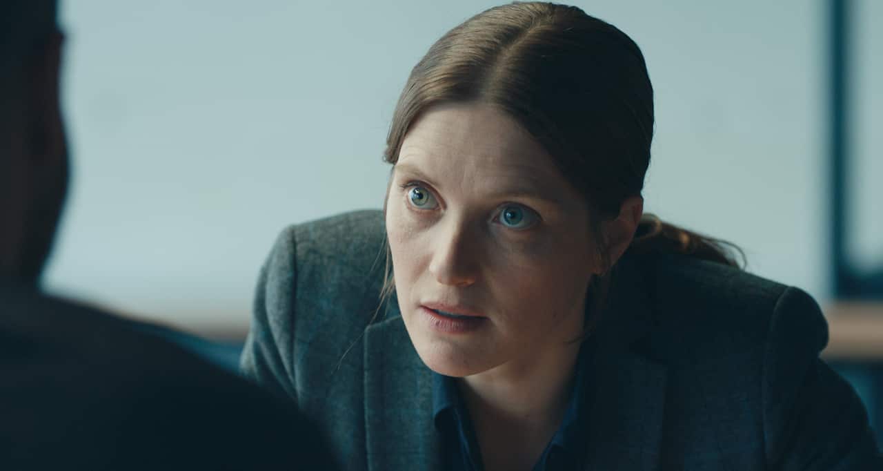 A woman in a grey jacket leans forward, as if talking to a person in front of her. 