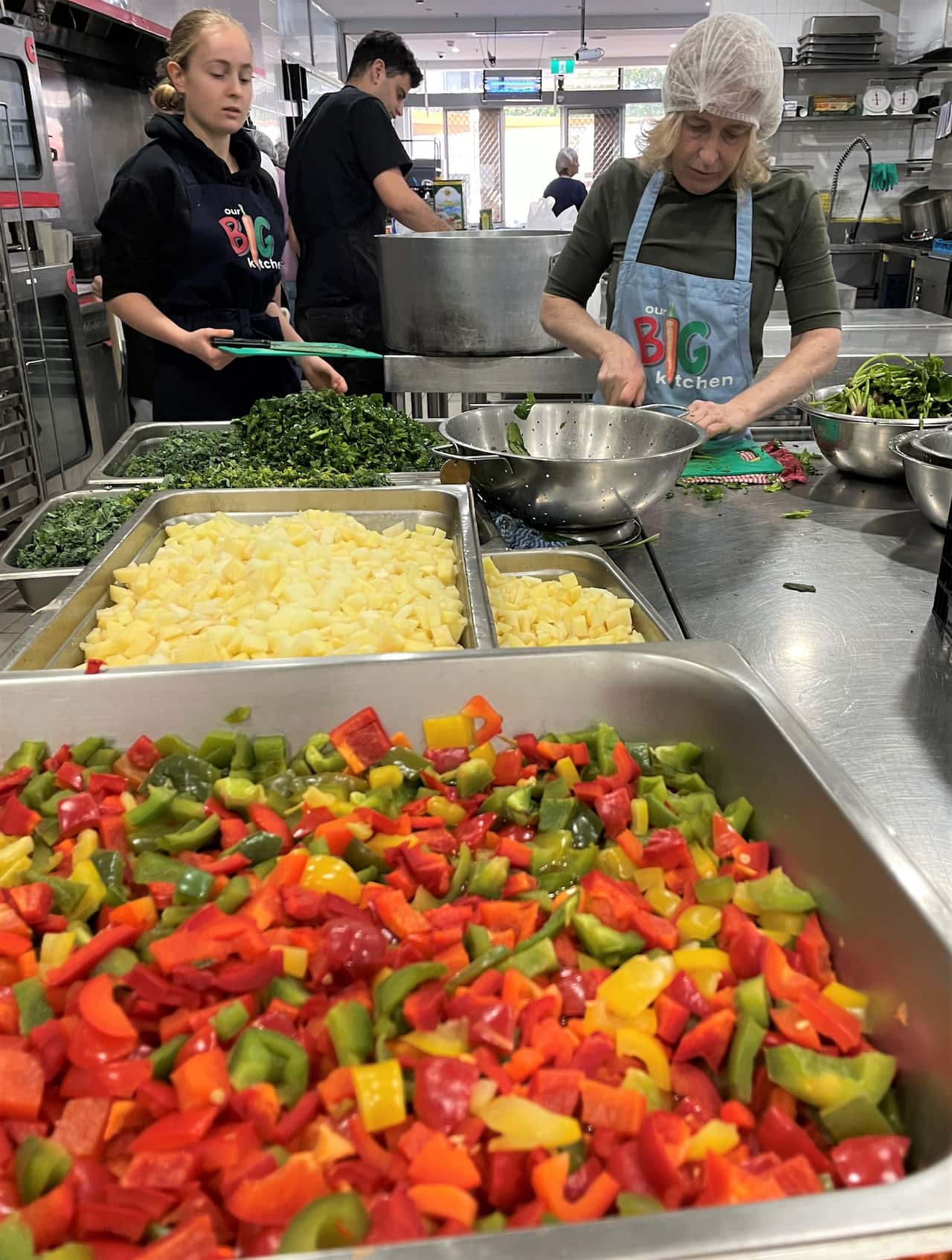 Two women stand behind a stainless steel counter behind tubs of colourful diced vegetables.