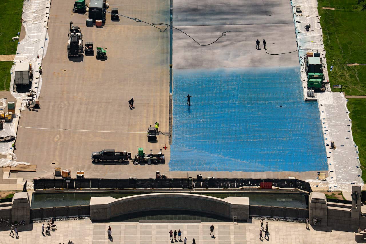 A drained Reflecting Pool seen from above with workers on its bare surface. One half of the pool base is blue.