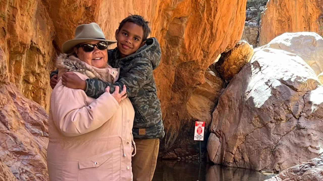 a middle aged woman in pink jacket, glasses and hat hugging a young boy in a camo jacket standing in front of orange rocks in the desert