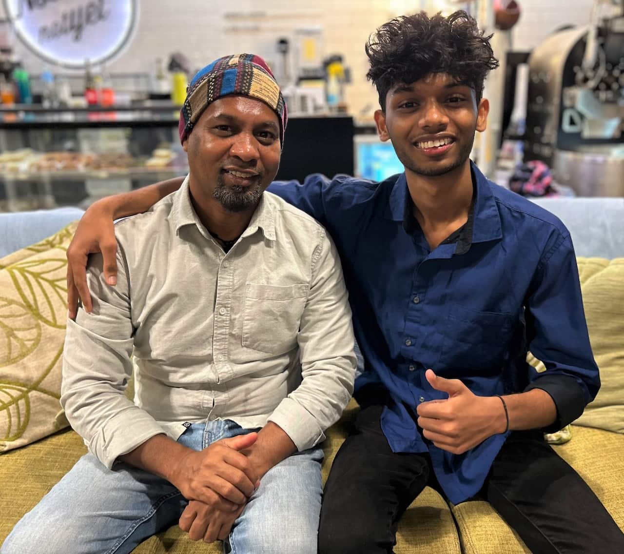Two men sit on a sofa, one wearng a grey shirt and the other a navy blue shirt.