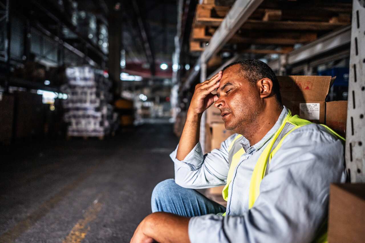 A worker in a warehouse, with his eyes closed and a hand to his forehead.