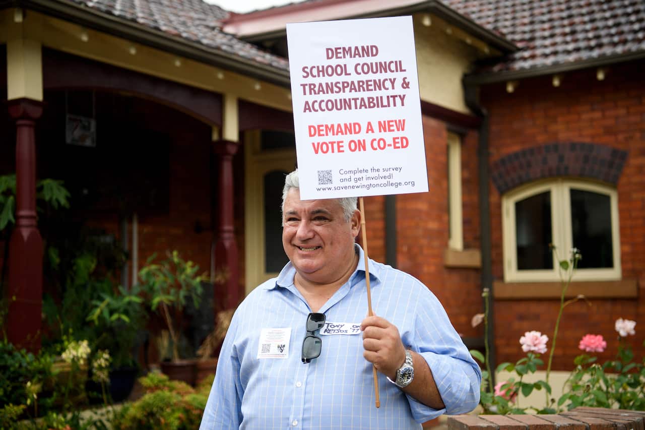 A man wearing a blue-checked shirt stands outside while holding a sign. 