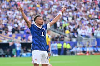 Kylian Mbappe, a French football player, dressed in blue and white with the number 10 on his chest, stands on a football pitch with his arms raised in celebration.