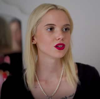 A portrait photo of a young woman with shoulder-length blonde hair, red lipstick, a pearl necklace and a serious expression on her face.