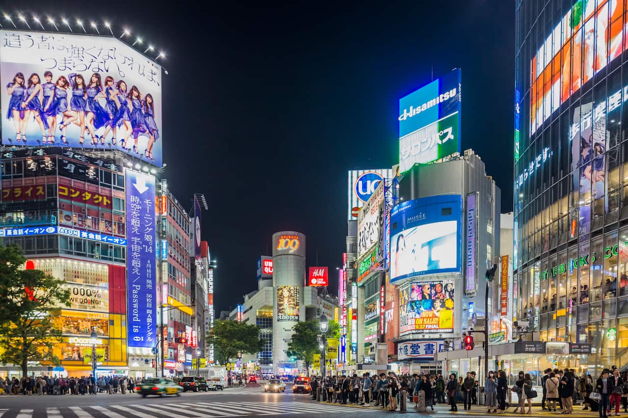 A street crossing lined with buildings displaying advertisements on screens, with a crowd walking through the area.