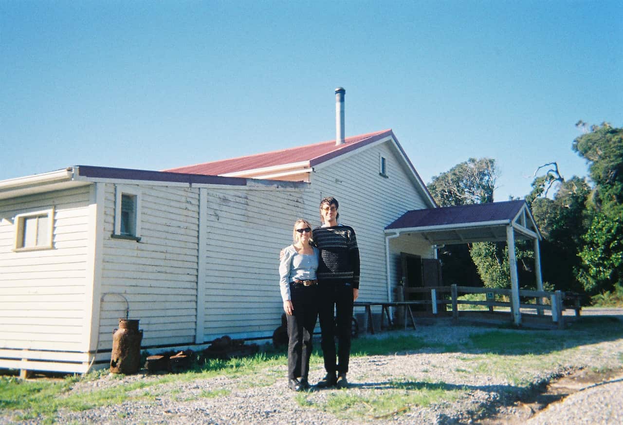 Two people pose next to a white weatherboard house.
