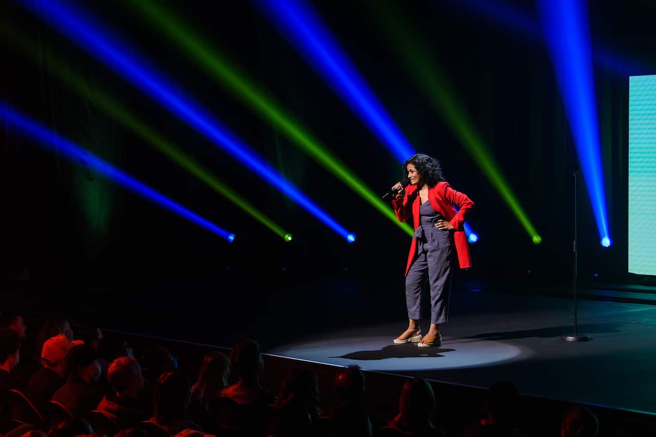A woman in grey pants and top and a red jacket stands on stage before an audience and talks into a microphone