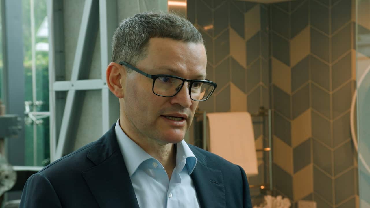 A man wearing prescription glasses and a dark suit with a light blue shirt underneath is speaking