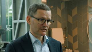 A man wearing prescription glasses and a dark suit with a light blue shirt underneath is speaking