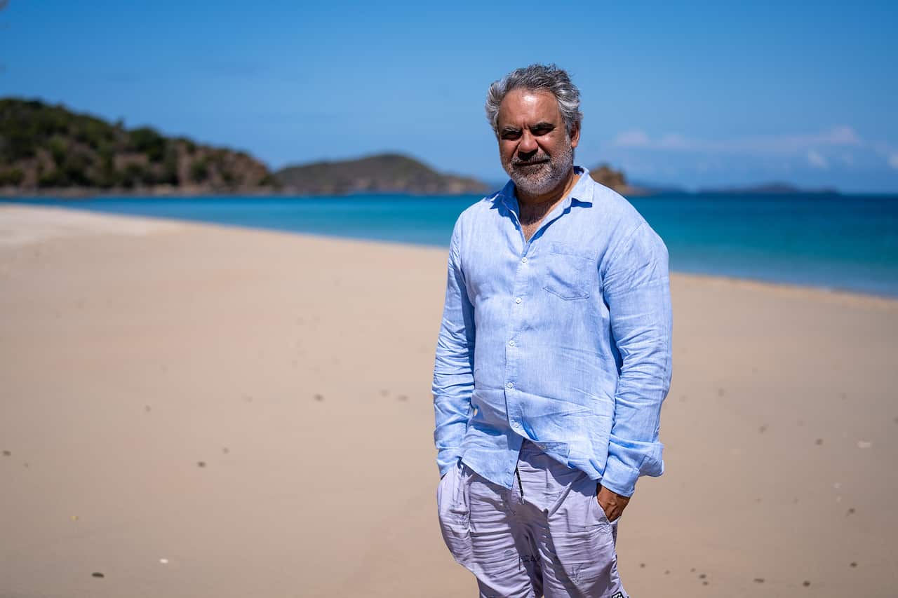 A man in a pale blue shirt stands on a beach, with the sea and coastline behind him.
