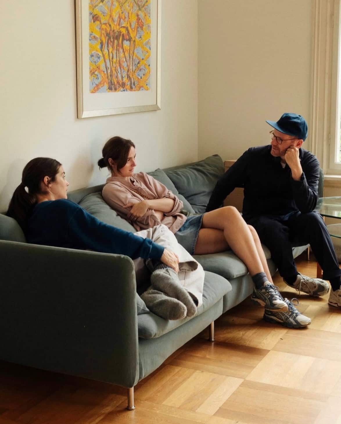 Three people sit together on a grey sofa in a brightly lit room, engaged in conversation beneath a framed piece of abstract yellow artwork.