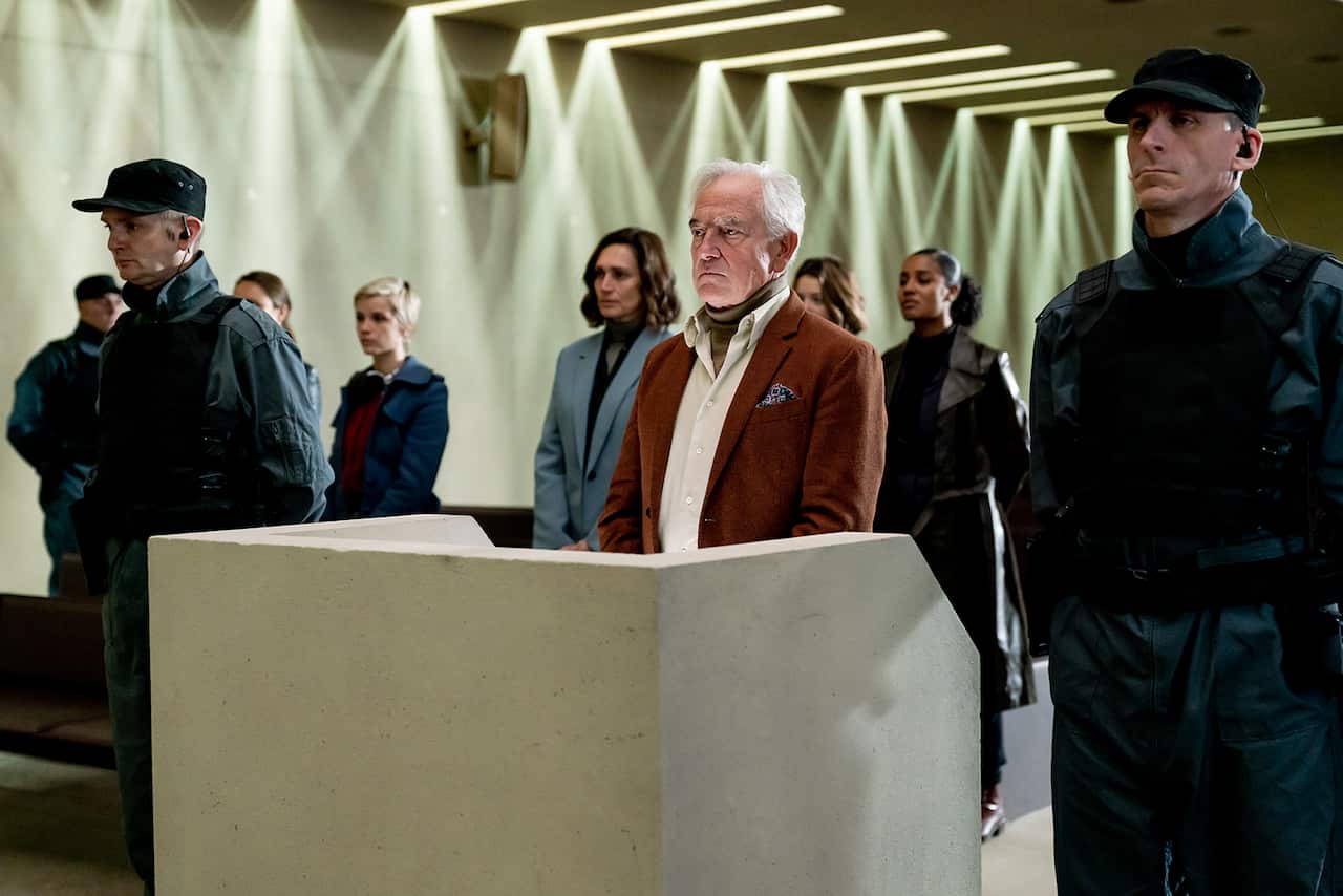 A man stands behind a concrete dock, withmen in black uniforms flanking him on both sides. Five women can be seen standing behind him. 
