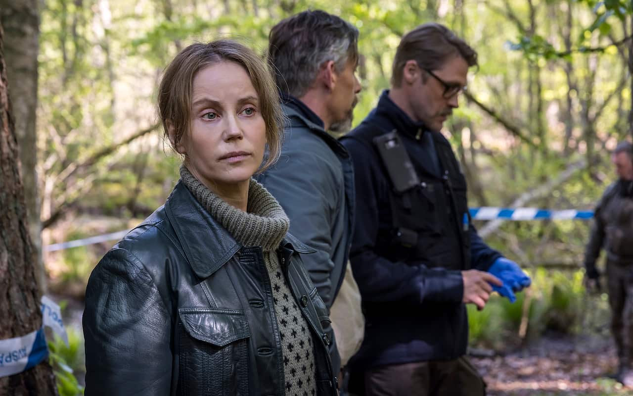 Seen had-and-shoulders, a woman in a turtleneck jumer and leather jacket stands in front of a bush scene, with crime scene tape running between trees behind her. Several men can be seen behind her.