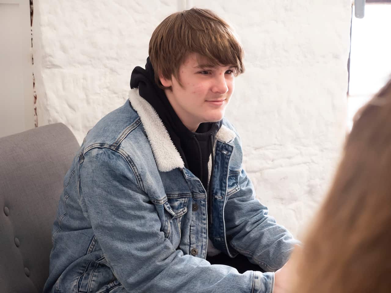 A teenage boy wearing a denim jacket and sitting in a chair