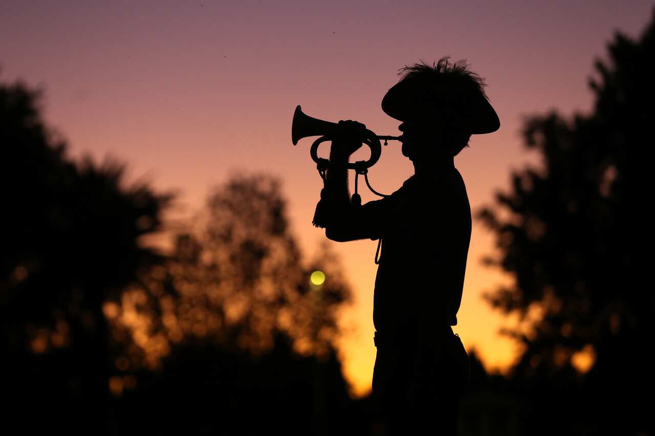 The silhouette of a man wearing a hat, playing a bugle at sunrise.