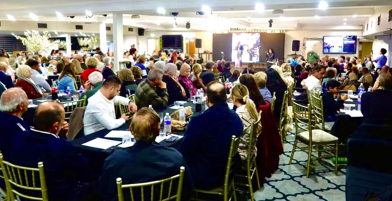A wide-angle photograph of a large, diverse group of people looking towards a person speaking at a podium.