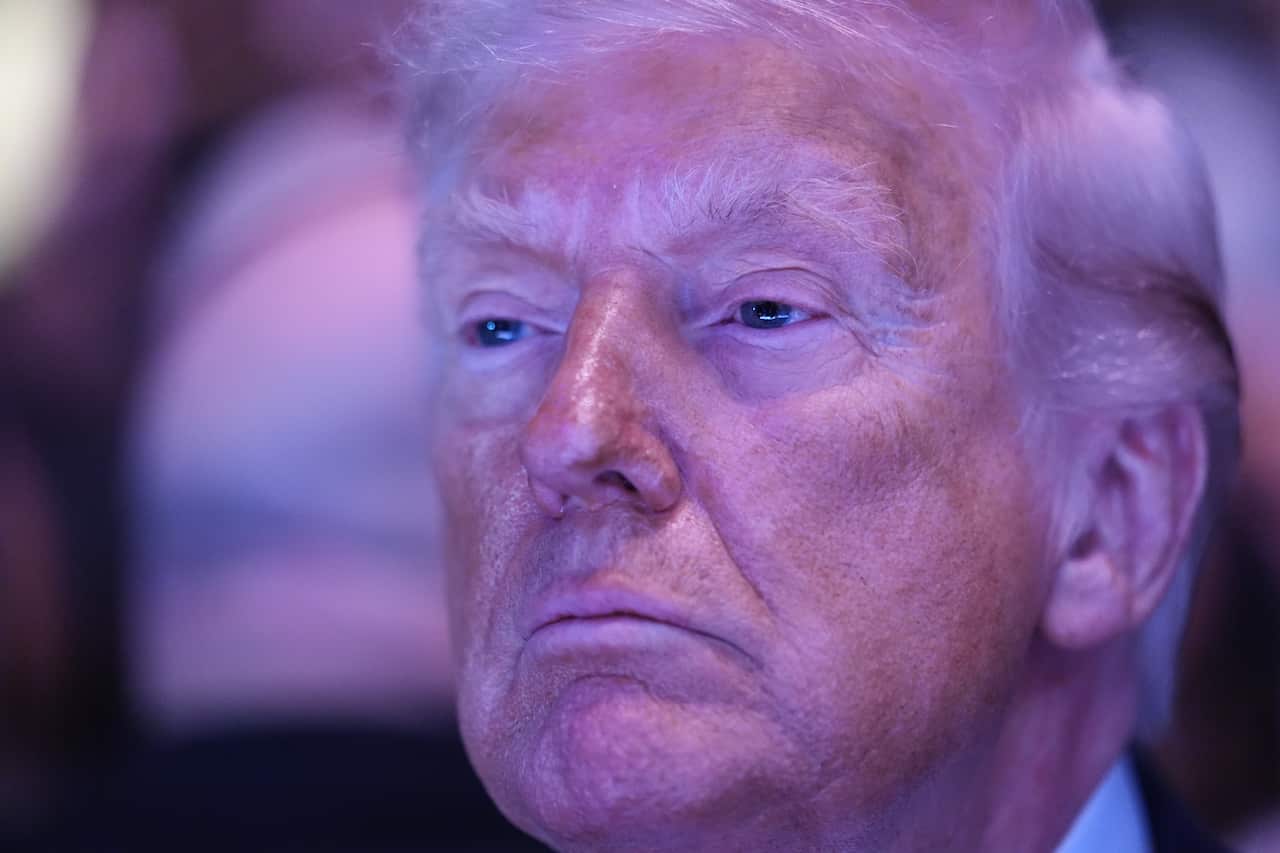 A close up of US president Donald Trump, cast in purple lighting. His expression is quite blank, impassive.