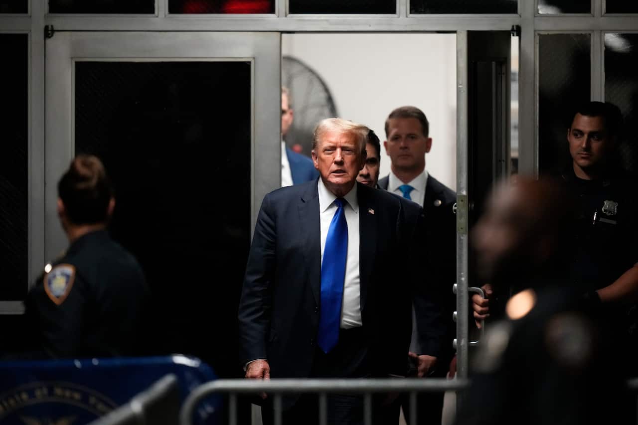 Donald Trump, wearing a dark suit and black tie, walks through a doorway into a courtroom.