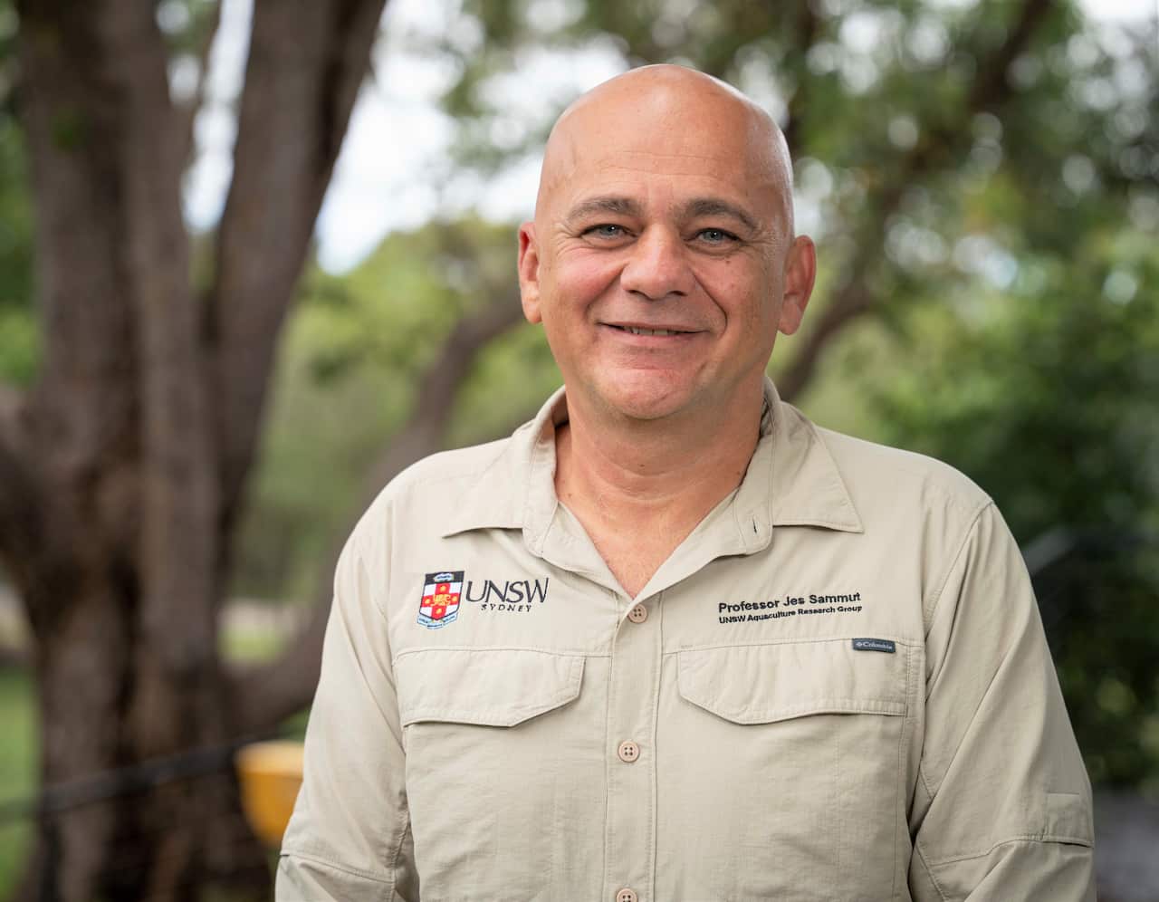 A man in a khaki shirt stands on a balcony in front of a green tree background. 