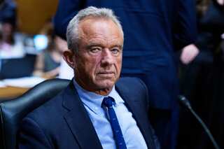 RFK Jr looks grim in a blue suit with speckled blue tie while sitting in a commitee