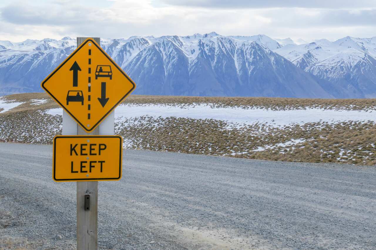 A 'keep left' sign next to a road with snow capped mountains in the background.