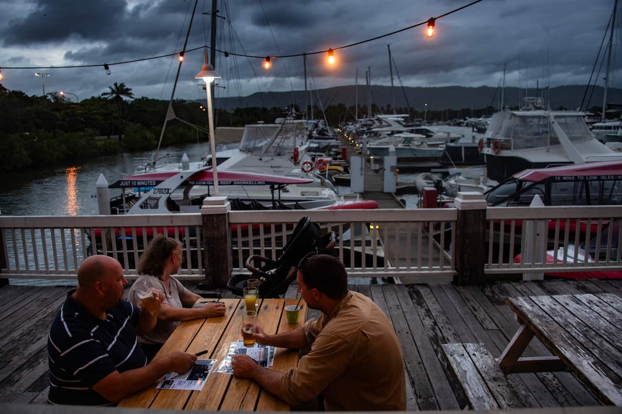 Four people sit at a wooden picnic table on a dock at dusk, enjoying drinks and conversation under a string of glowing lights with a marina full of sailboats and yachts in the background.