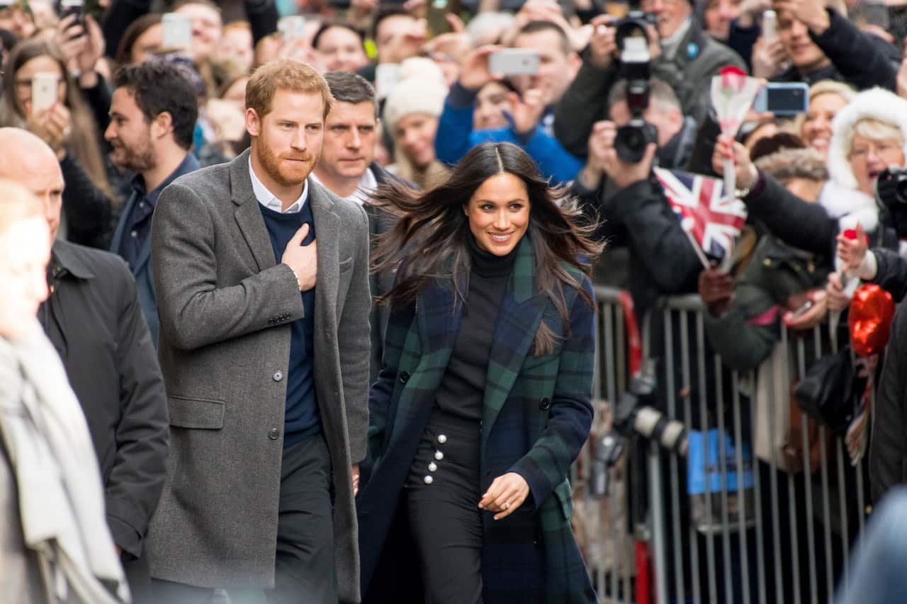 Prince Harry and his wife hold hands as they walk in public. A large crowd is in the background.