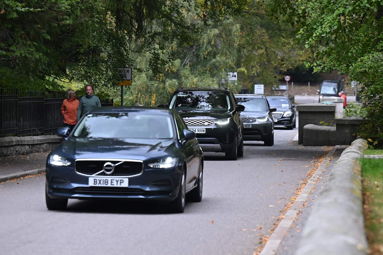 A convoy of dark-colored vehicles drives down a road lined with trees and a stone wall. In the foreground, a dark blue Volvo leads the procession, with two people walking on a paved path to the left of the cars.