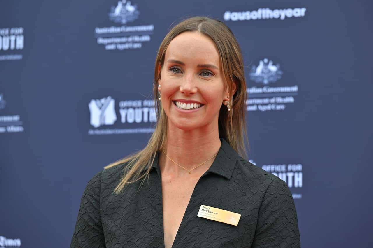 A brunette woman wearing a black dress poses in front of a media wall