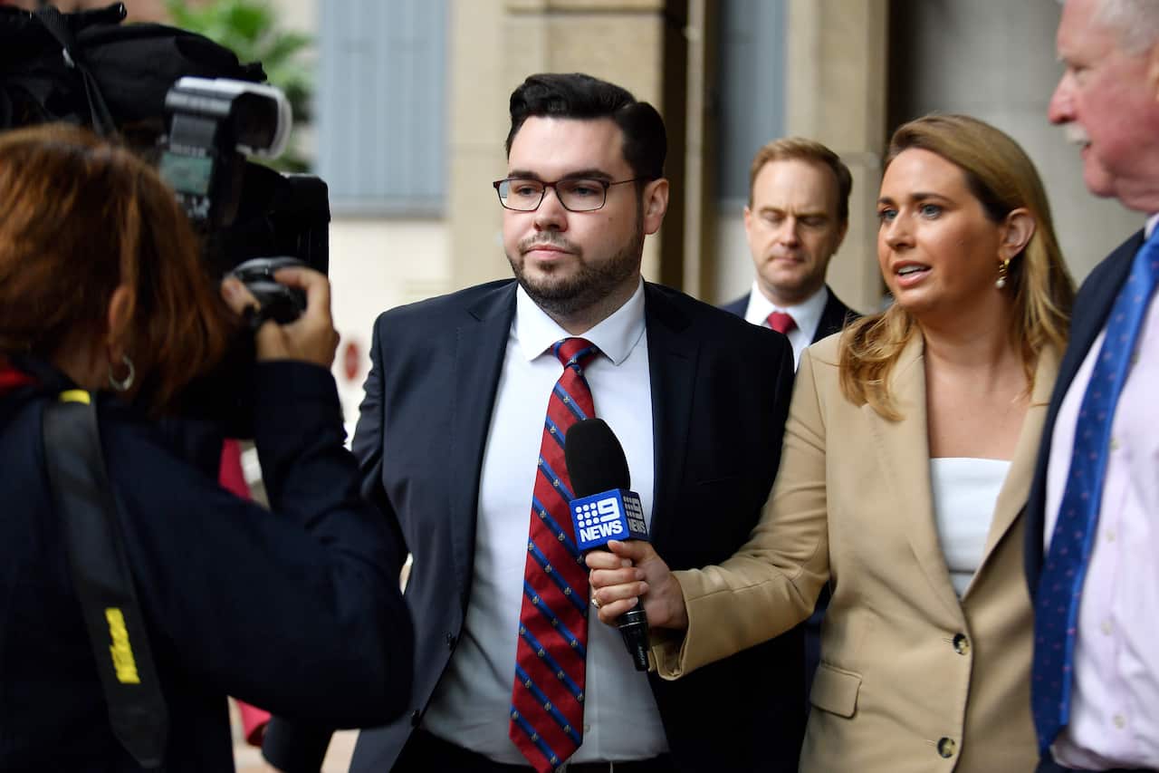 A man wearing a suit, tie, and glasses walks as a journalist holds a microphone in front of him.