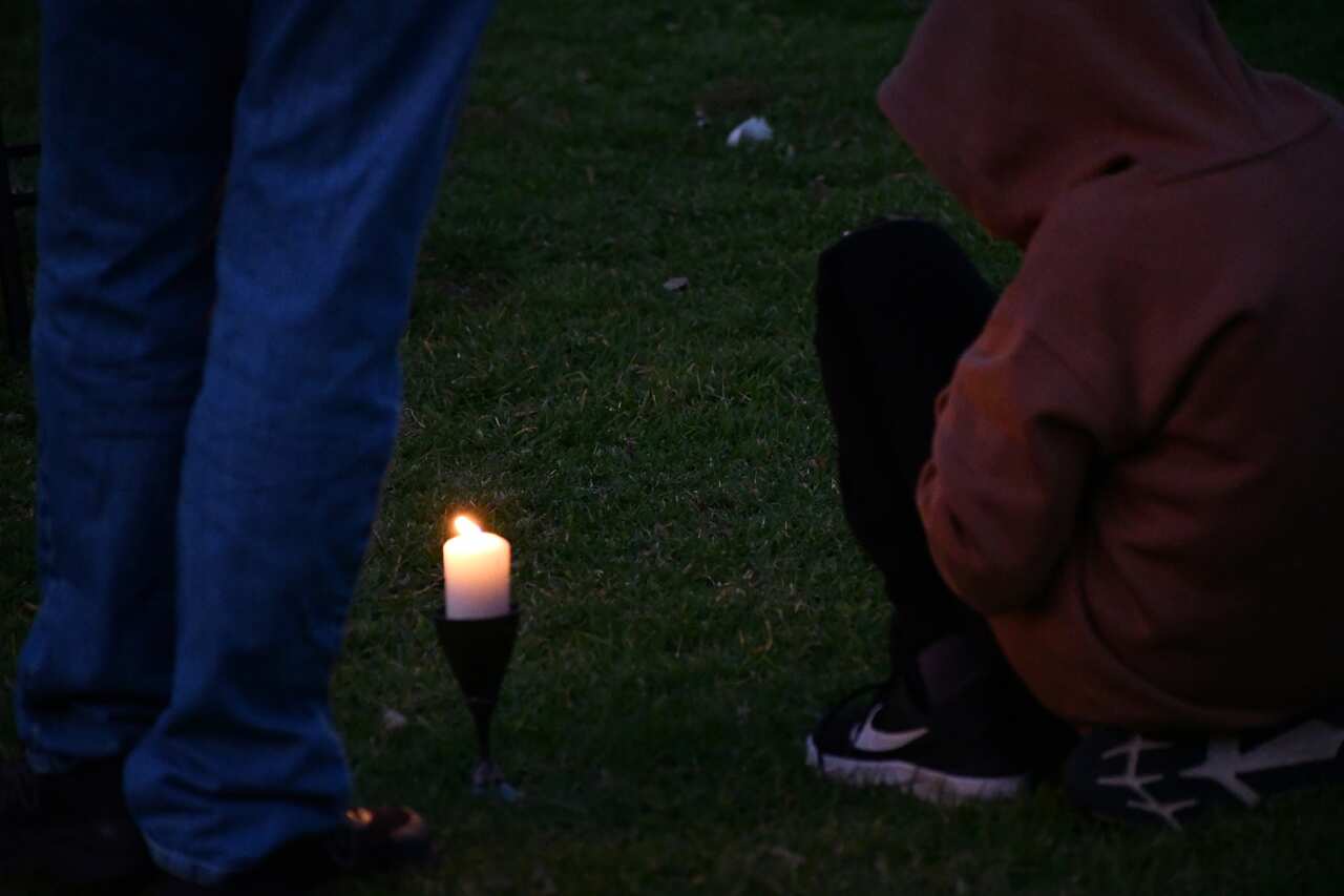 People sit and stand near a lit candle on a field.