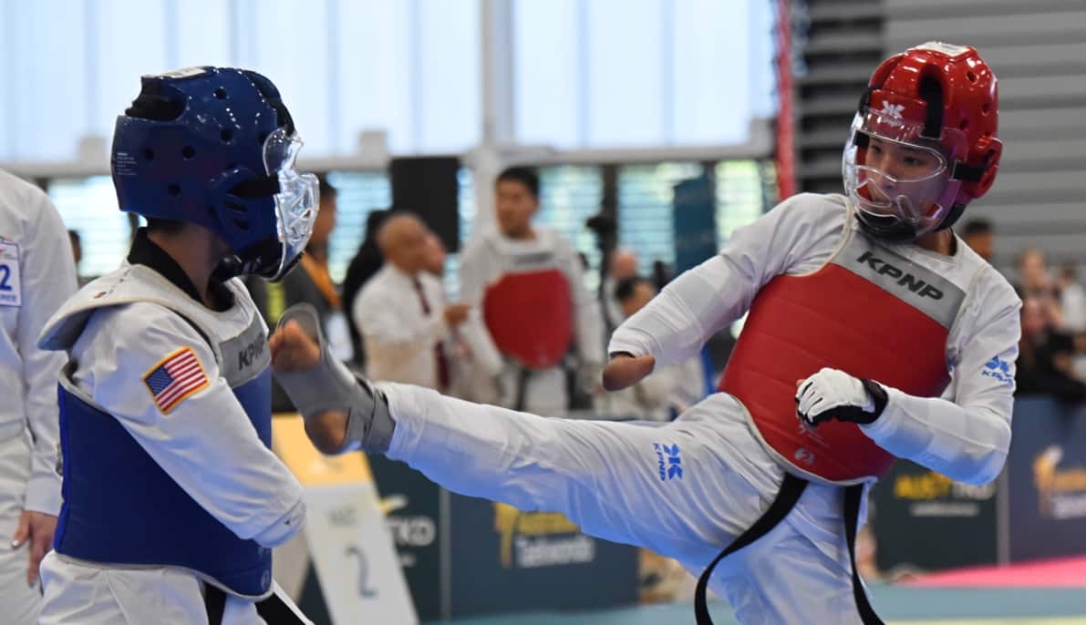 Two athletes engaged in taekwondo. One is wearing a blue vest and helmet, and the other is wearing a red vest and helmet.